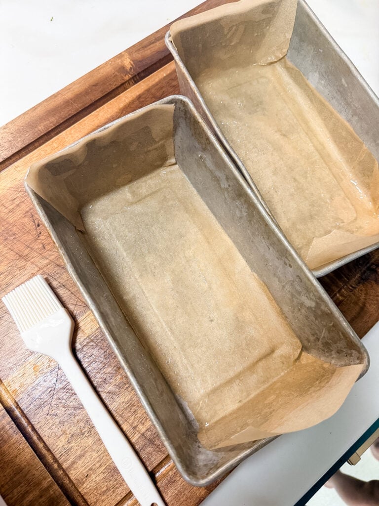 Two empty greased loaf pans lined with parchment paper on a wooden cutting board