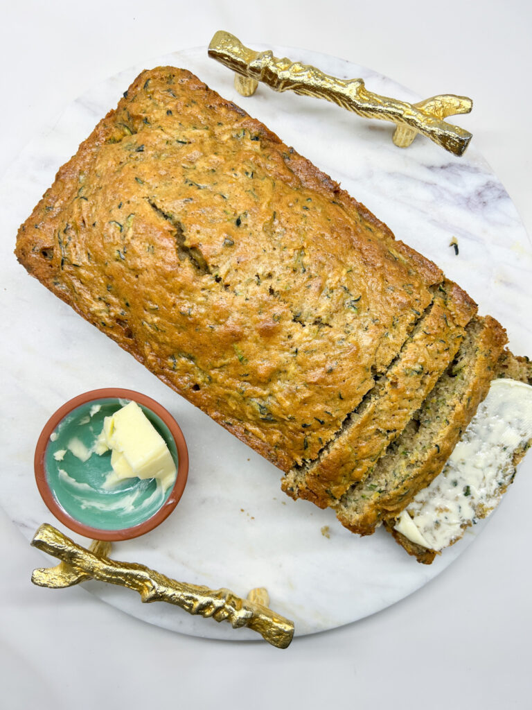 Two golden brown zucchini loaves on a white marble board with a small teal dish of butter and gold branch decor