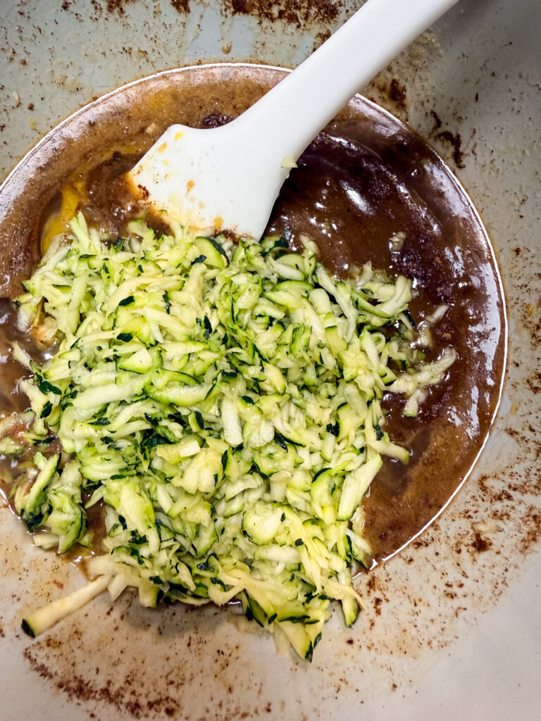 White silicone spatula folding bright green shredded zucchini into brown batter in a speckled mixing bowl