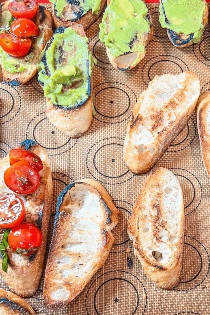 several slices of bread on a baking sheet some with avocado and some with tomatoes