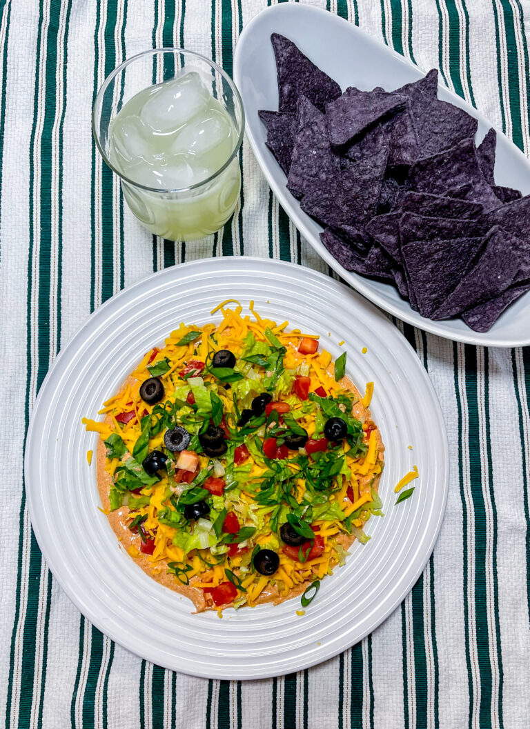 Finished taco dip next to a bowl of blue corn chips a glass of a margarita on ice