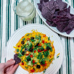 Finished taco dip next to a bowl of blue corn chips a glass of a margarita on ice and a hand dipping a chip into the dip.