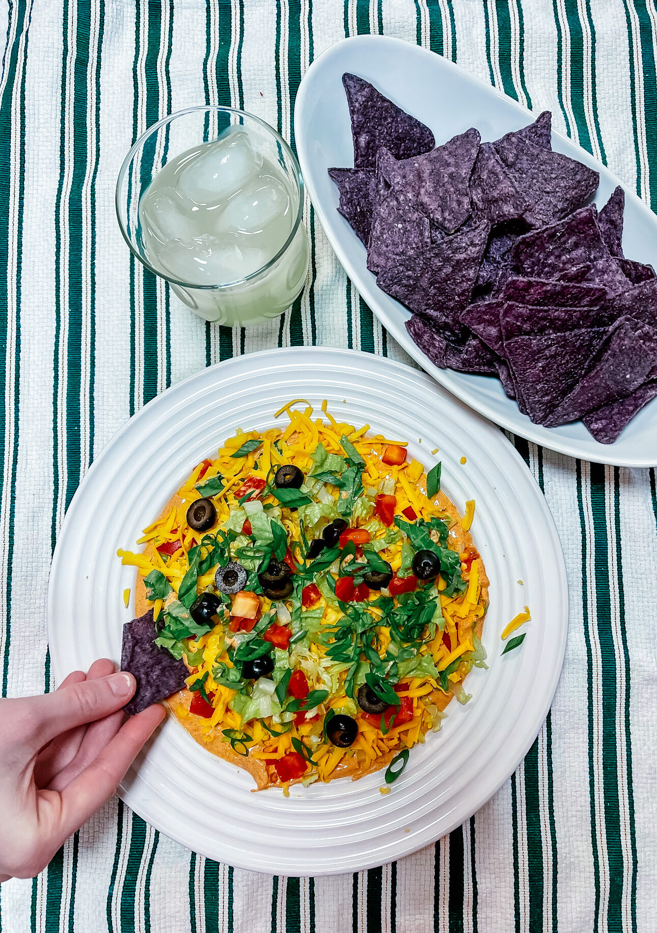 Finished taco dip next to a bowl of blue corn chips a glass of a margarita on ice and a hand dipping a chip into the dip.
