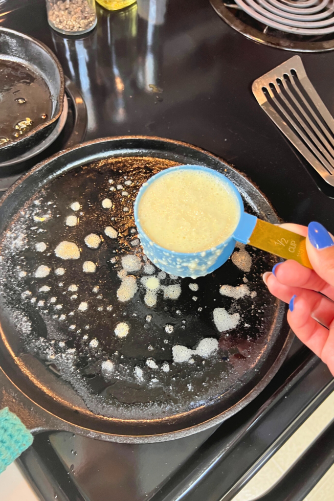 a person holding a measuring cup of pancake batter over a pan