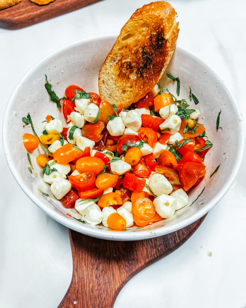 Finished Marinated Cherry Tomato and Mozzarella Salad in a ceramic bowl with a crostini in the salad. The bowl is on top of half of a cuddle board set.
