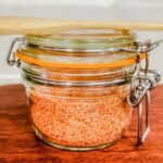 My Everything bbq rub in a jar on a wood board with a bamboo spoon on top.