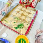 One of my girls sprinkling Sanding Sugar onto the cut sugar cookies on a baking sheet.