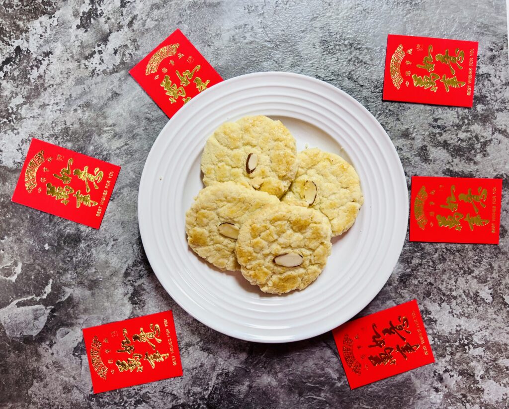 Finished Chinese almond cookies on a white circle plate surrounded by Chinese red envelopes, on a dark gray marble counter.