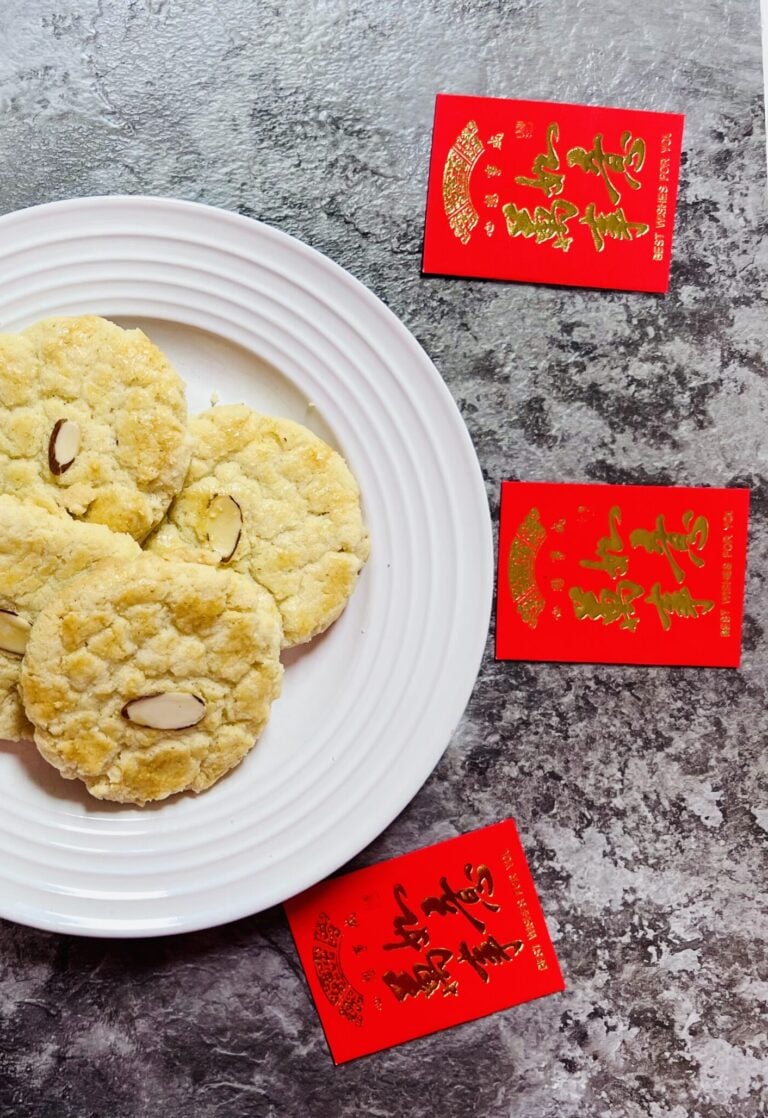 Finished Chinese almond cookies on a white circle plate surrounded by Chinese red envelopes, on a dark gray marble counter.
