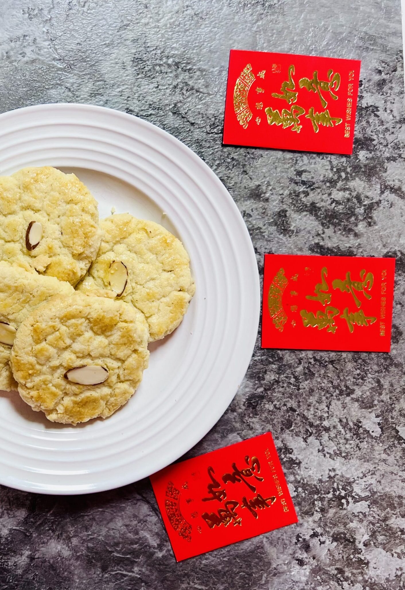 Finished Chinese almond cookies on a white circle plate surrounded by Chinese red envelopes, on a dark gray marble counter.