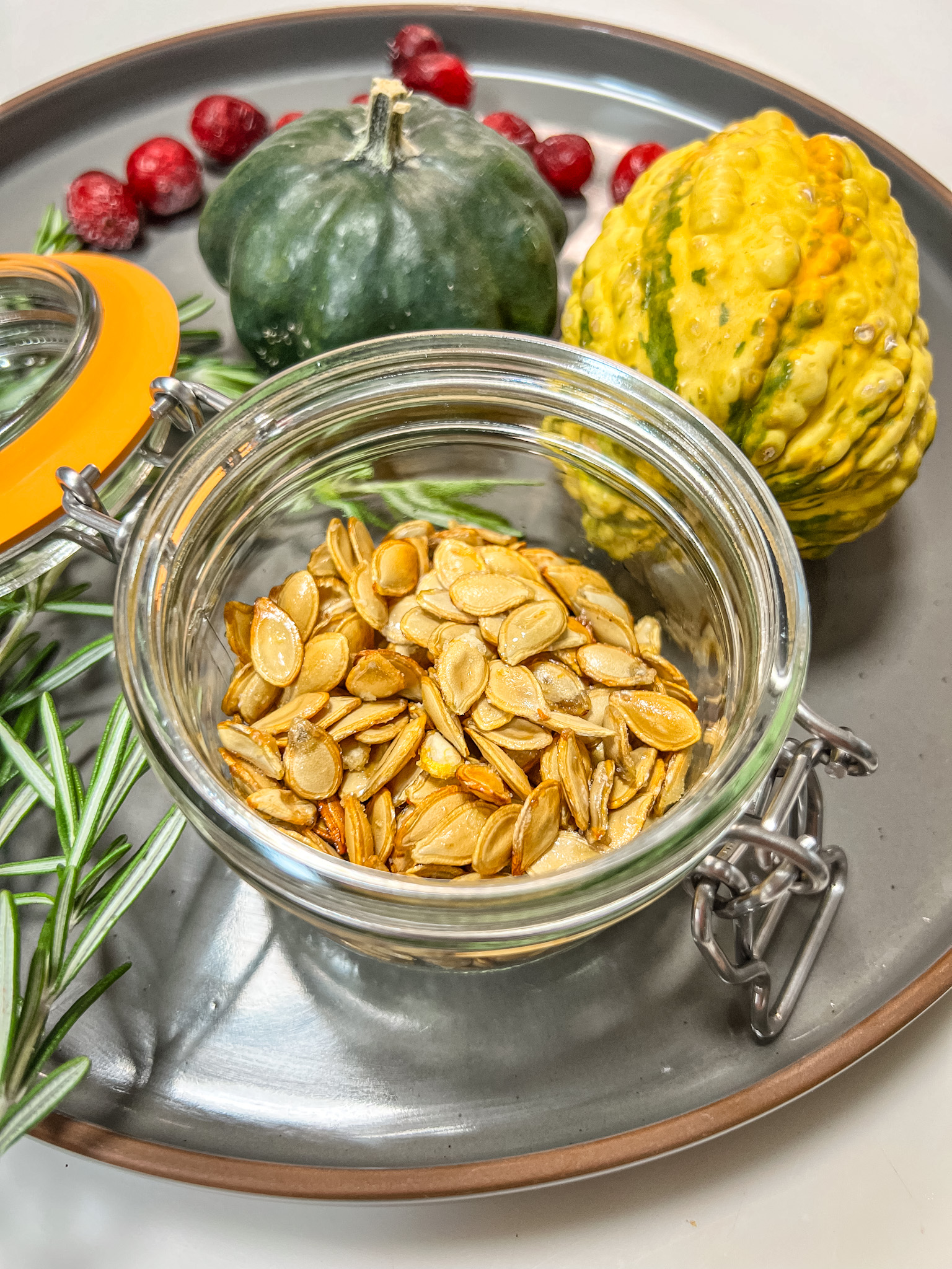 Roasted quash seeds sitting in a glass jar on a ceramic plate surrounded by mini gourds, cranberries, and rosemary sprigs.