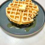 waffle on a ceramic plate with a slab of butter top and syrup.