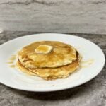 Image of pancakes with a small pad of butter, and maple syrup on a white plate.