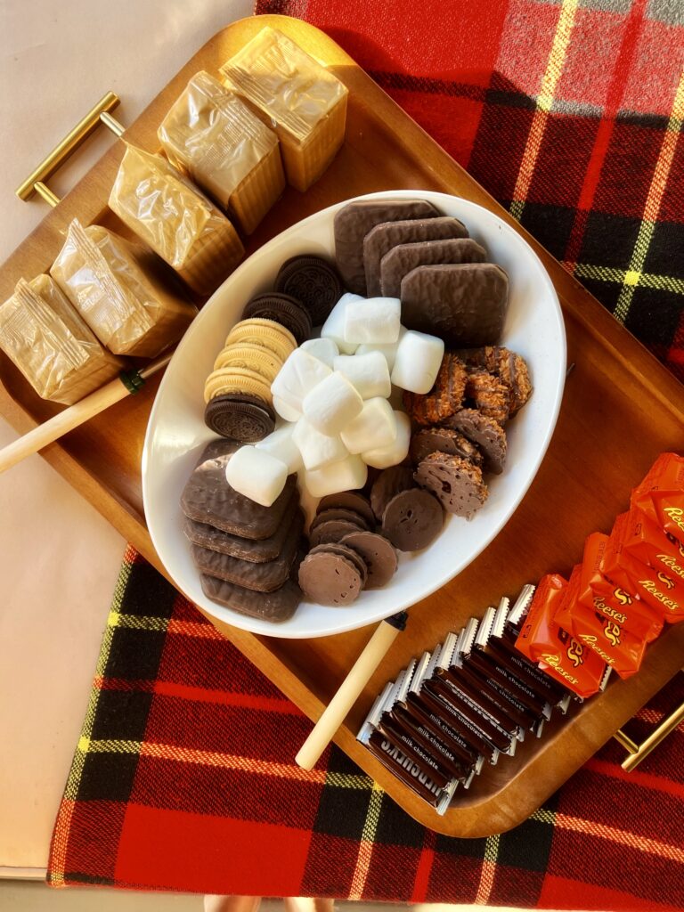 A wood tray with graham crackers, skewers/marshmallow forks, a ceramic platter with cookies and marshmallows, and chocolate bars on the other side of the ceramic platter on the wood tray.