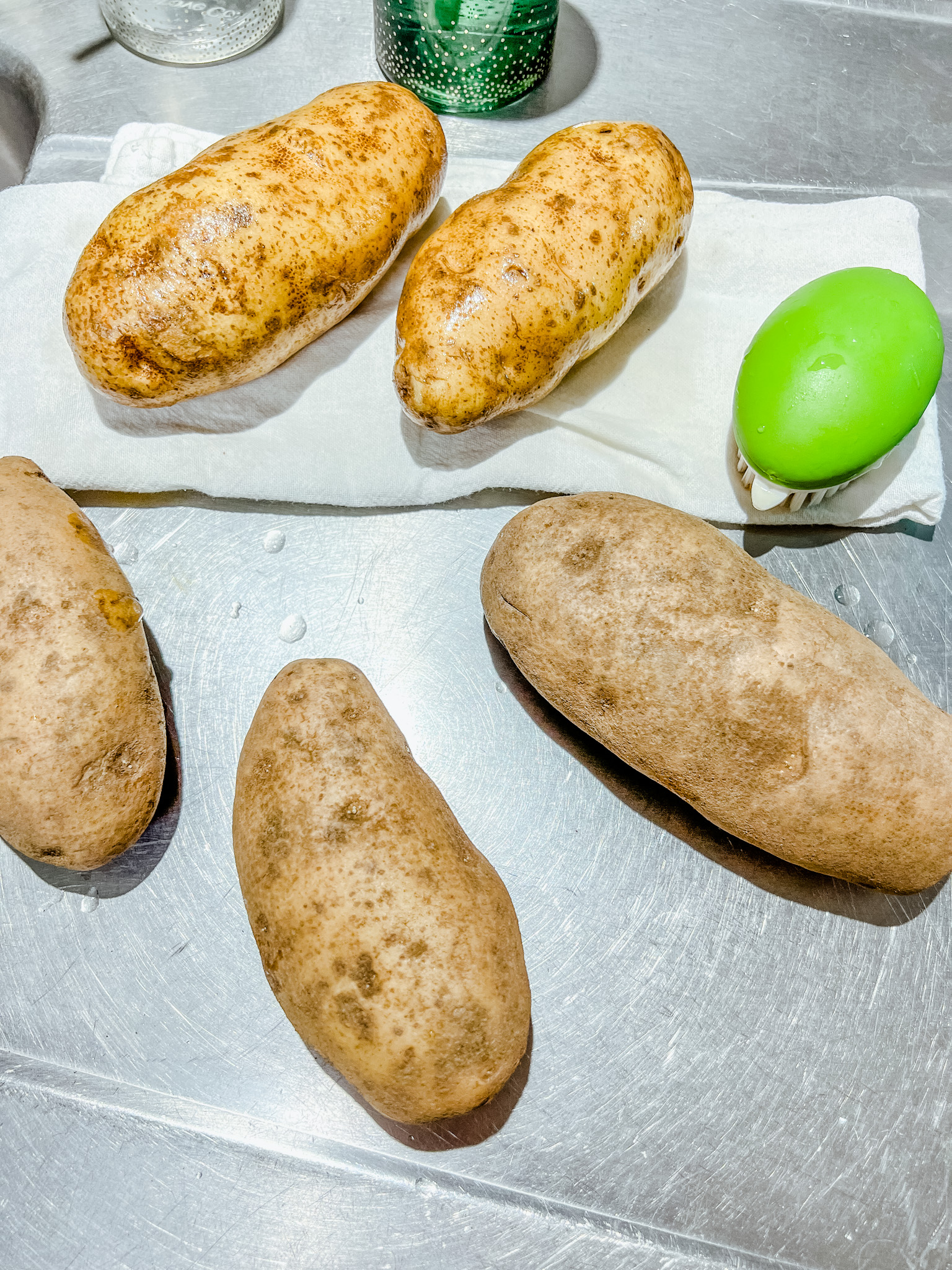 Clean potatoes on a towel next to a vegetable scrubber and some potatoes not cleaned yet.