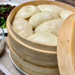 Header image. Cooked, bao buns in a bamboo steamer basket, the lid leaning against the basket