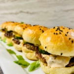 Close up image of Pork Wonton Burgers in a line on a platter with homemade black sesame seed buns and asian cucumber salad