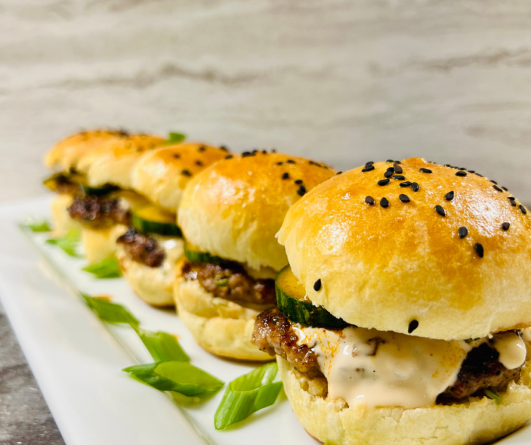 Close up image of Pork Wonton Burgers in a line on a platter with homemade black sesame seed buns and asian cucumber salad