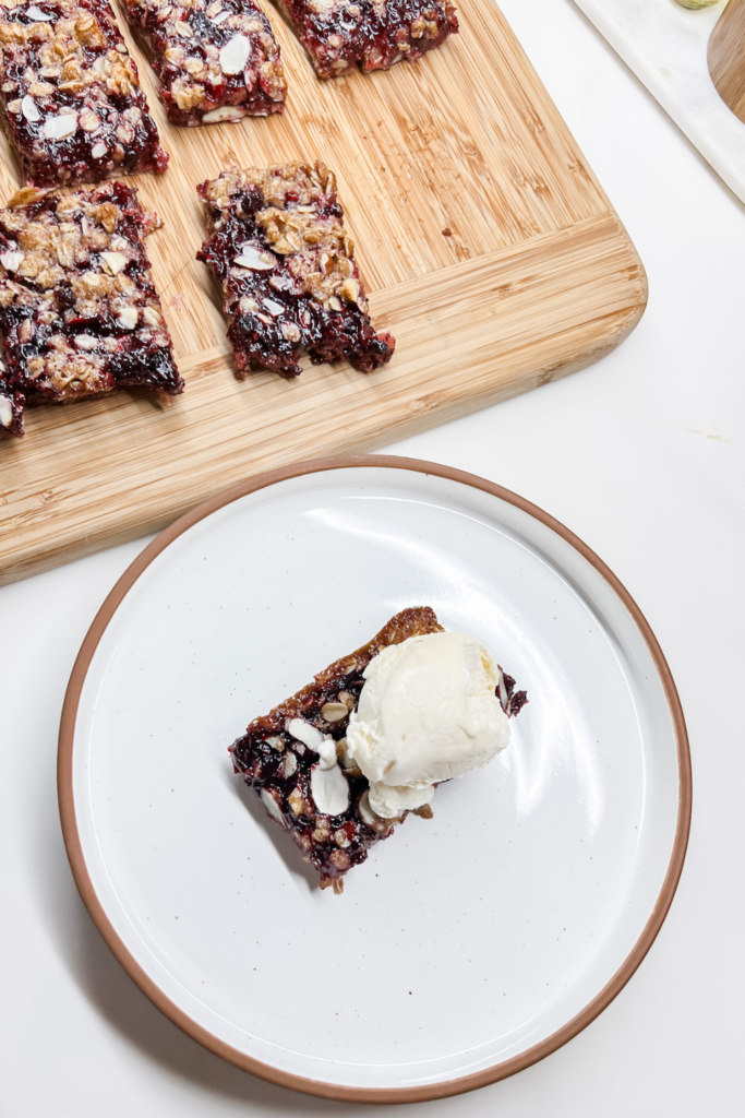 Finished Almond Berry Bar with ice cream on a white ceramic plate next to remaining bars on a bamboo cutting board.