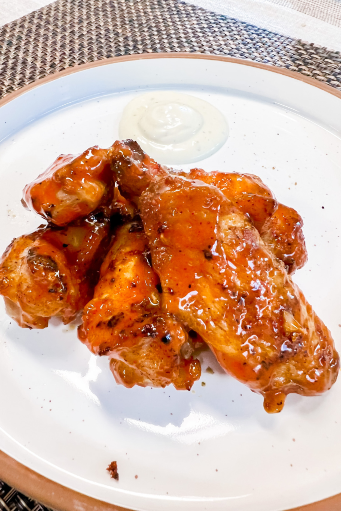 Finished grilled wings after being tossed and covered in Buffalo Sauce on a white ceramic plate with a dollop of ranch in the background.