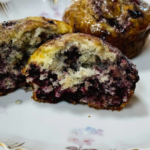 Blackberry orange muffins image finished muffins one cut in half to show all the berries in side. The muffin is sitting on a white porcelain plate.