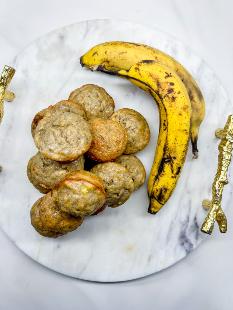 a pile of mini banana muffins with bananas laying next to the pile. all on a marble planner