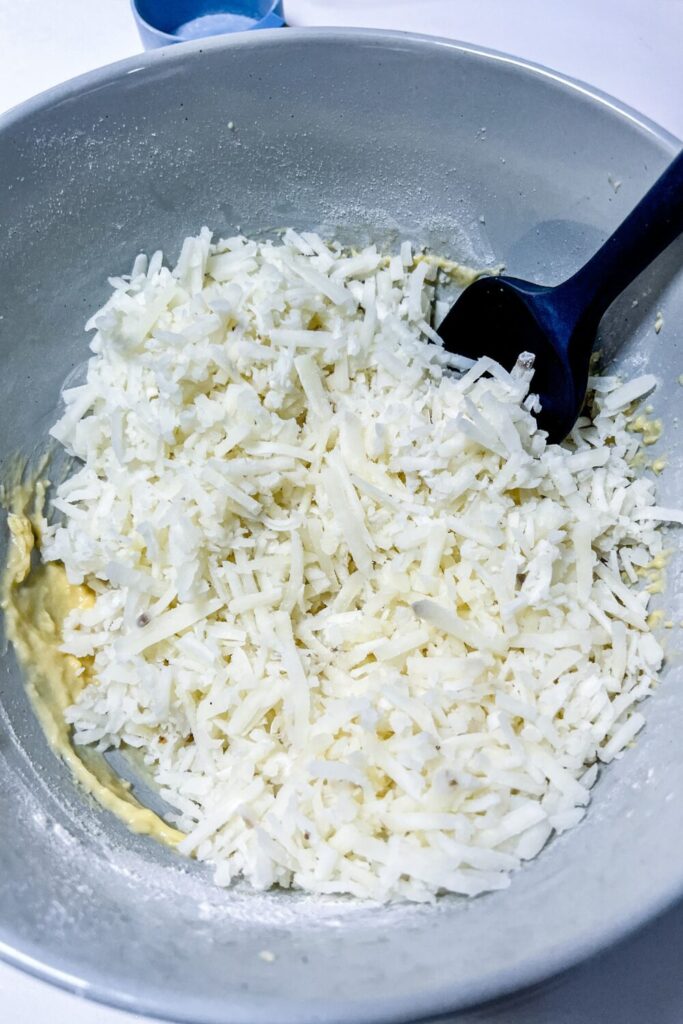 a bowl of defrosted hashbrowns on top of the potato pancake batter with a spatula in it