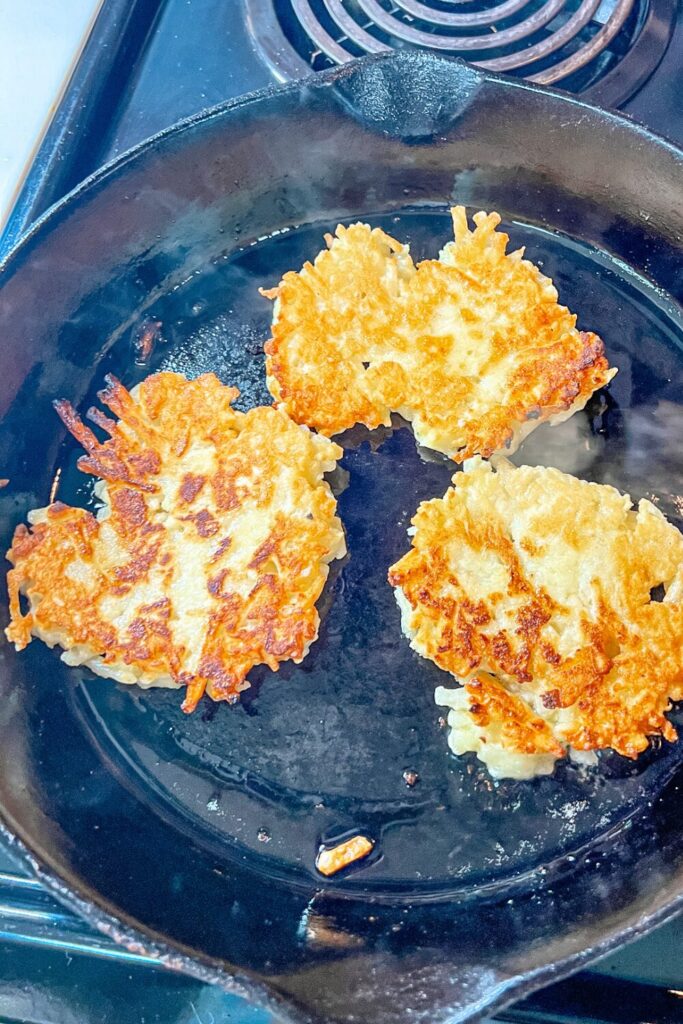 potato pancakes cooking in a skillet on the stove after being flipped (side shown is golden brown)
