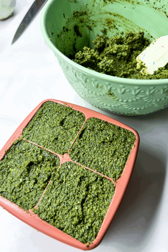 a bowl of green pesto with a spatula next to a silicone portion tray filled with pesto for freezing