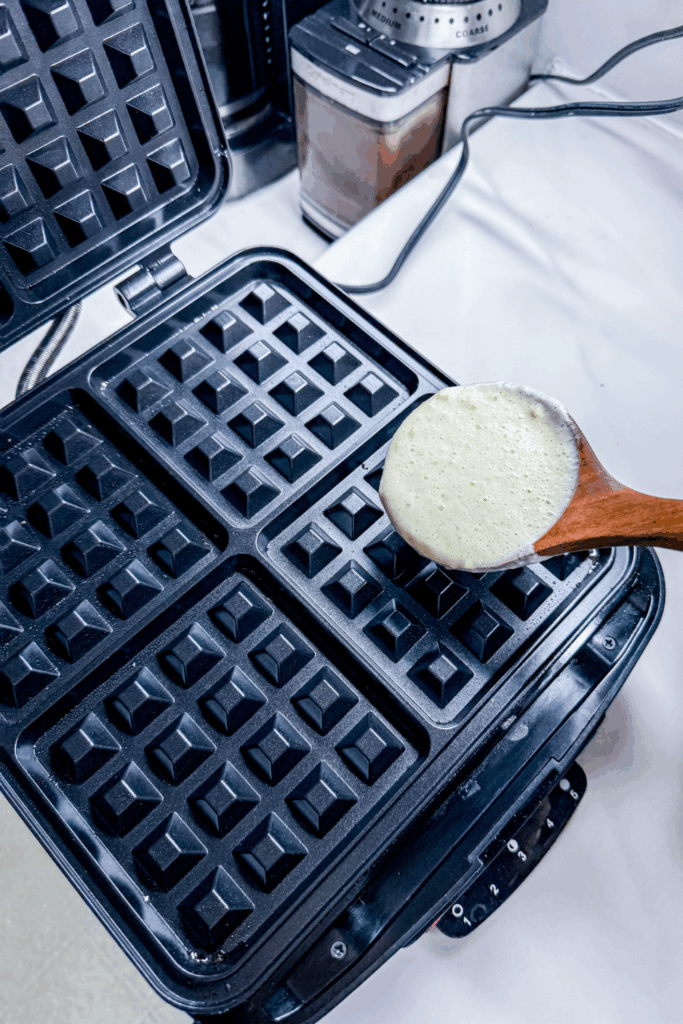 A waffle iron is being used to make waffles. A laddle of batter is above one well ready to fill