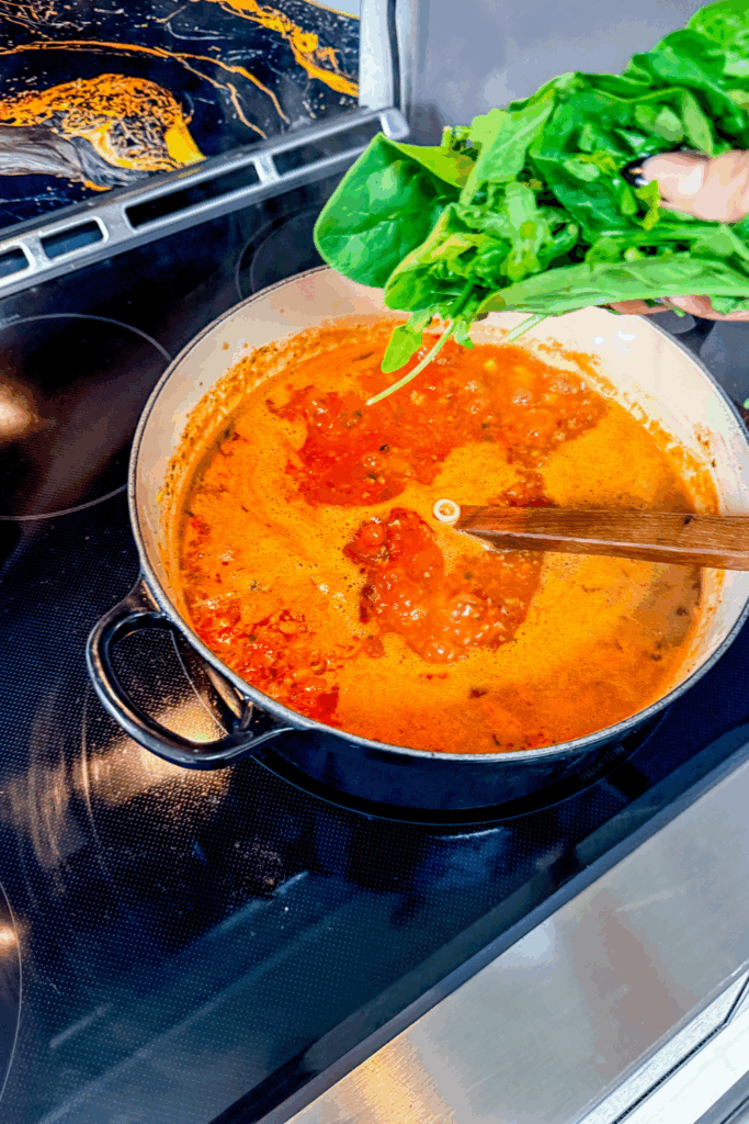 adding the spinach and arugula to the A person holding a measuring spoon over a pot of the Italian Ground Sausage Soup