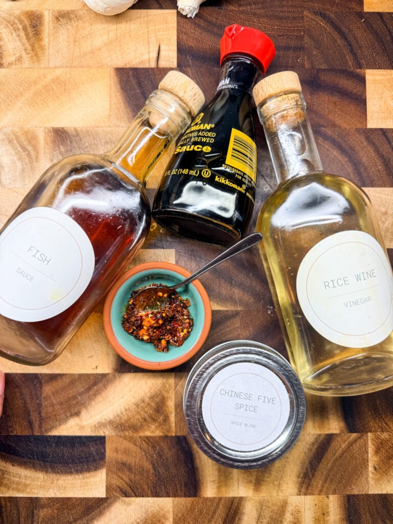 Asian pantry staples including soy sauce, fish sauce, rice wine vinegar, chili crisp, and Chinese five spice on a wooden cutting board