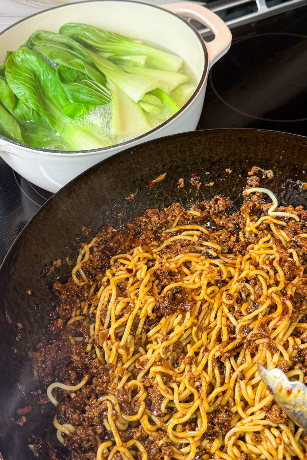 Blanching bok choy in boiling water while Dan Dan noodle sauce cooks in a wok