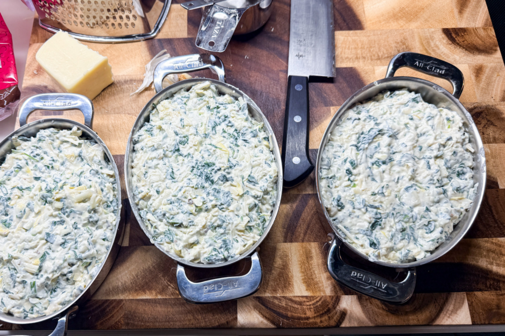 Spinach artichoke dip mixture divided into three small casserole dishes before adding shredded cheese topping.