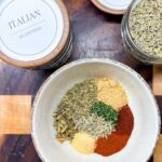 All the spices for homemade Italian sausage seasoning measured into a white bowl before mixing, with Italian seasoning and fennel seed jars on a wood cutting board.
