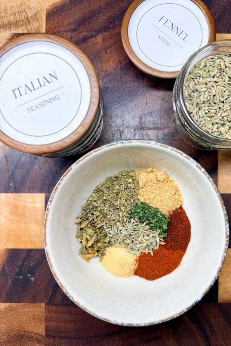 All the spices for homemade Italian sausage seasoning measured into a white bowl before mixing, with Italian seasoning and fennel seed jars on a wood cutting board.