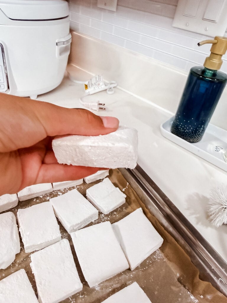 Hand holding a single thick homemade marshmallow square coated in powdered sugar, showing soft pillowy texture