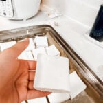 Hand holding a single homemade marshmallow square coated in powdered sugar above a tray of finished marshmallows