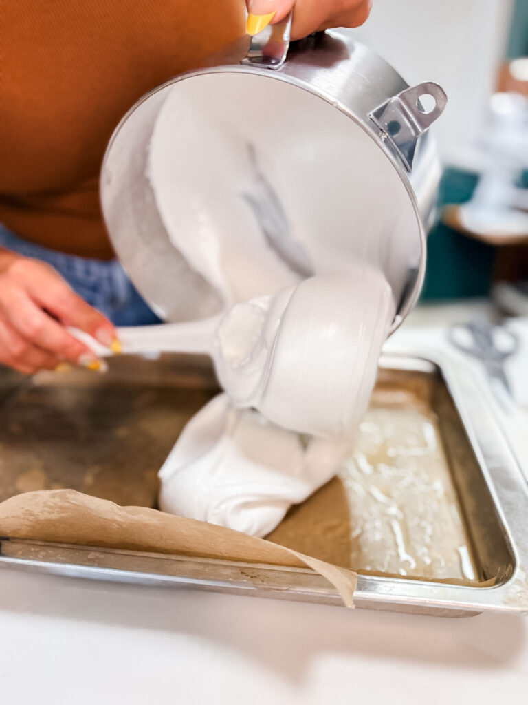 Marshmallow mixture falling in thick ribbons from a stand mixer bowl into a lined baking pan