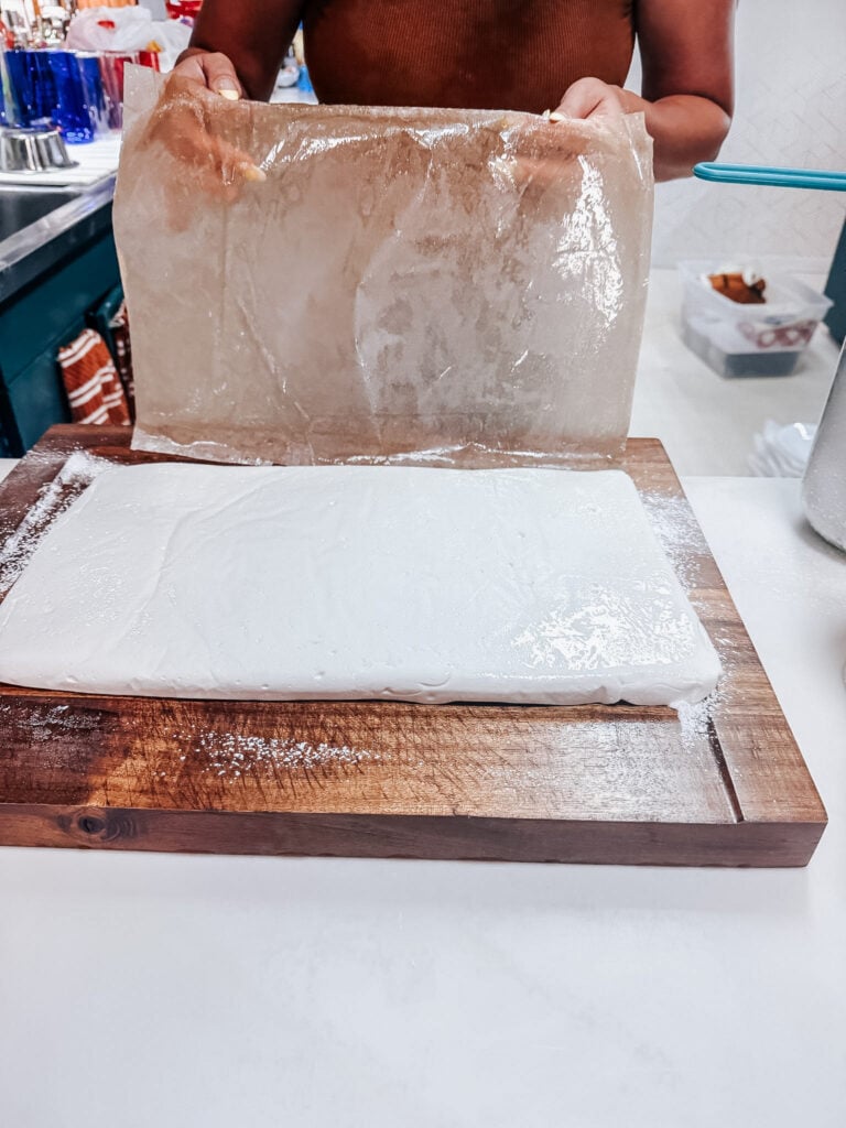 Peeling plastic wrap off the set marshmallow slab on a wooden cutting board dusted with powdered sugar