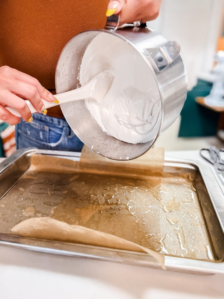 Pouring fluffy white marshmallow mixture from a stand mixer bowl into an oiled plastic wrap-lined baking pan