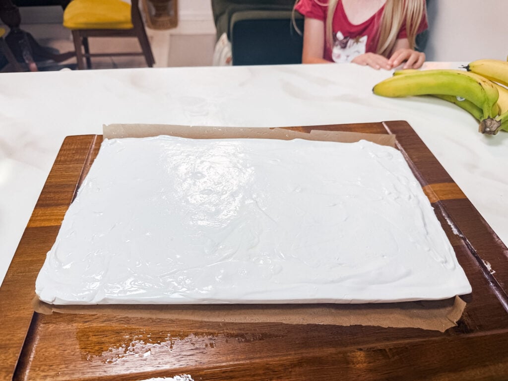 Flat white marshmallow slab resting on a wooden cutting board on a kitchen counter, with a child sitting in the background