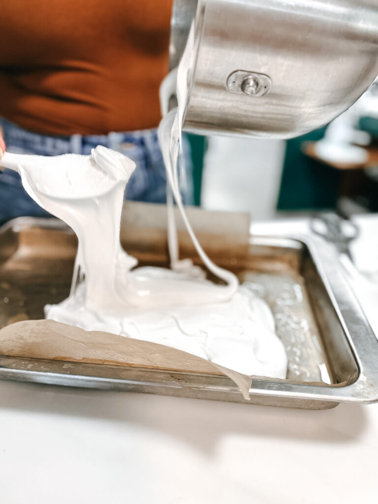 Hands using an offset spatula to spread white marshmallow mixture evenly in a baking pan lined with plastic wrap