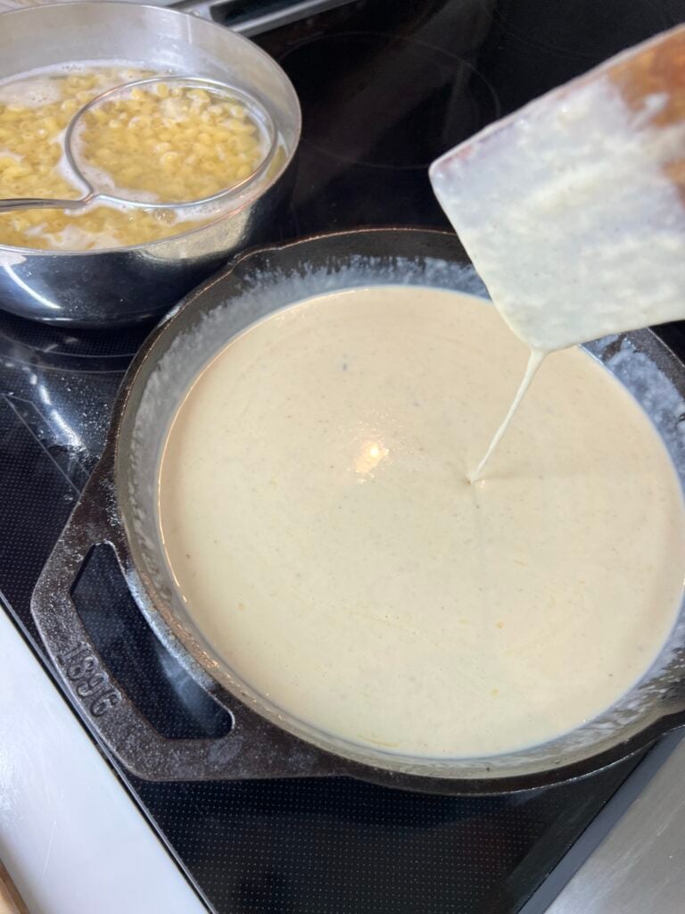 Creamy white roux-based cheese sauce being poured into a Lodge cast iron skillet, smooth and silky with steam rising, mac and cheese in a pot visible in the background