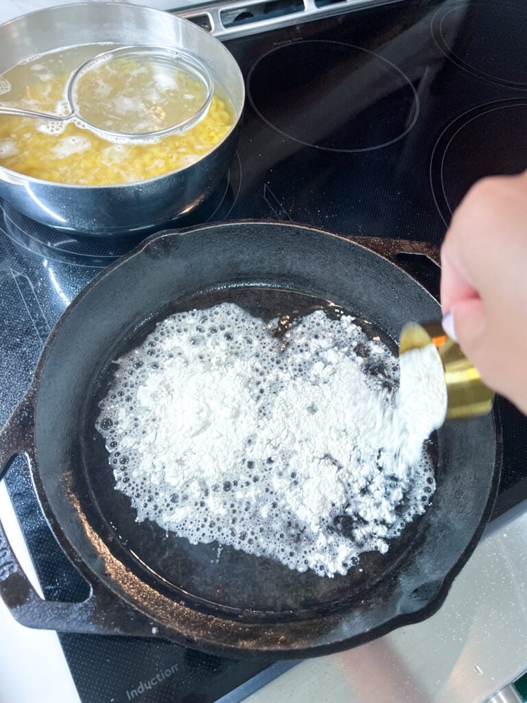 Overhead view of a smooth white roux being whisked in a seasoned cast iron skillet on an induction stovetop, bubbly and pale cream in color