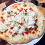Finished white pizza on a white plate with melted mozzarella, fresh burrata, diced tomatoes, basil, and seasoned chicken on a golden blistered crust