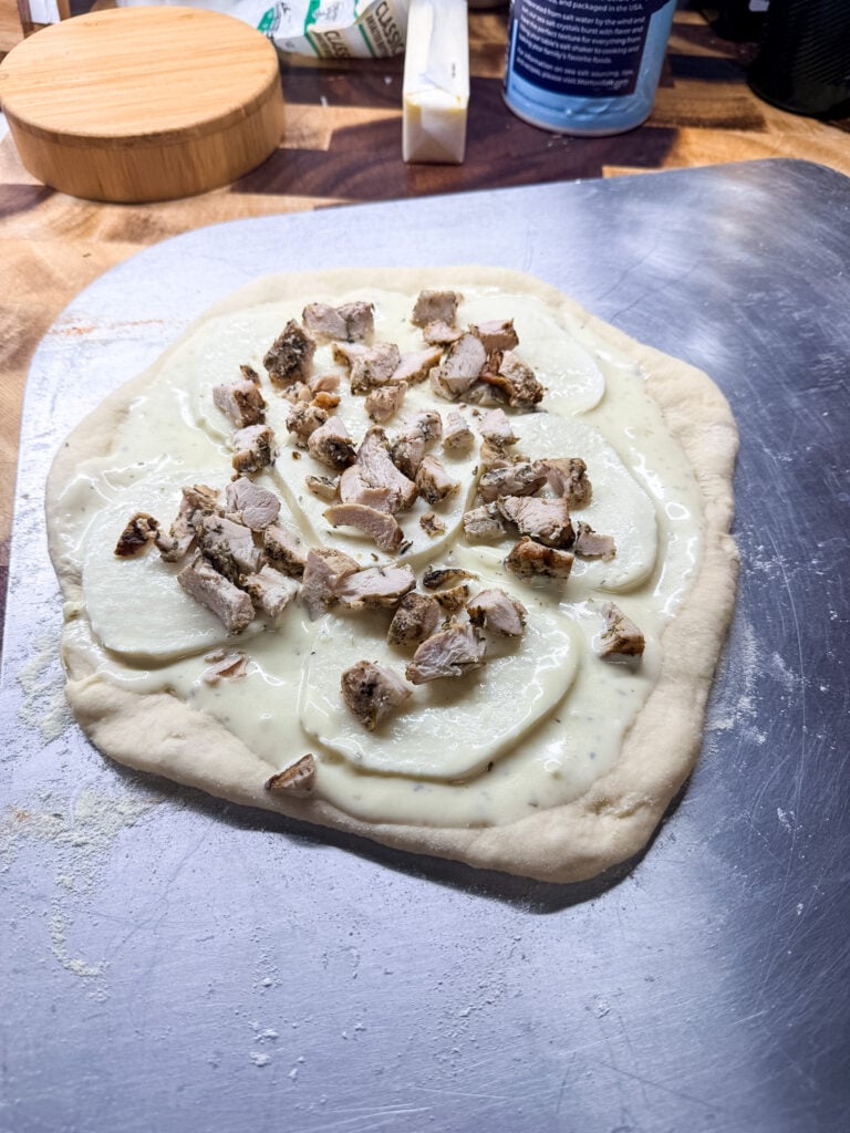 White pizza sauce spread over stretched homemade pizza dough on a metal pizza peel, topped with seasoned chopped chicken before baking