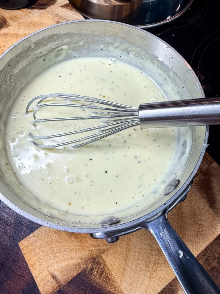 White pizza sauce being whisked in a stainless steel saucepan on an induction stovetop, showing smooth creamy texture with flecks of Italian seasoning