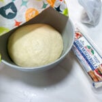 A ball of homemade pizza dough after rising in a gray bowl, covered with a colorful kitchen towel, with a package of fresh mozzarella nearby.