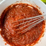 Homemade red pizza sauce being whisked in a white bowl with dried herbs
